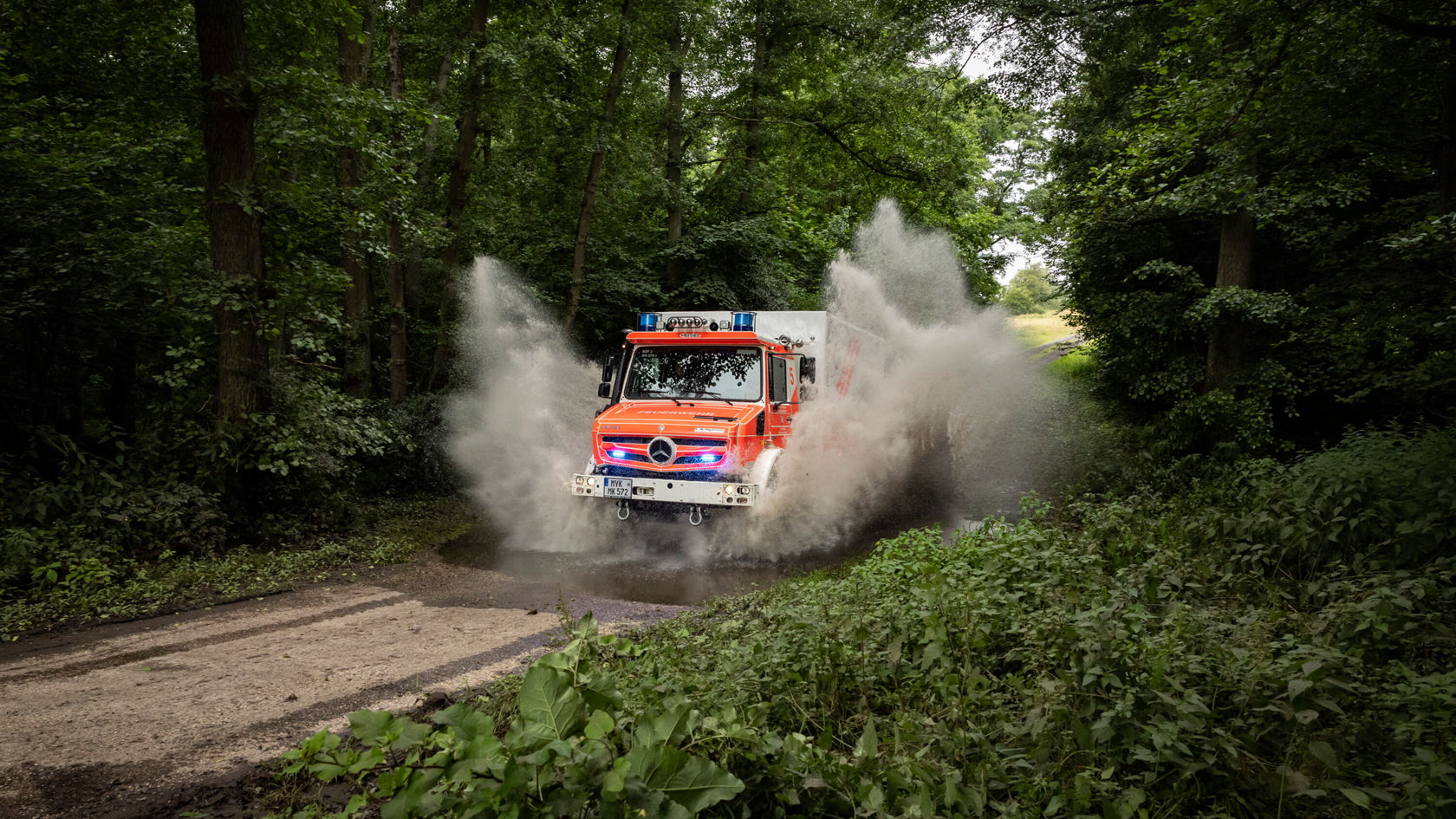Un Unimog Mercedes‑Benz traverse une flaque dans une forêt et projette de l'eau et de la boue tandis qu'il circule sur un chemin non goudronné.