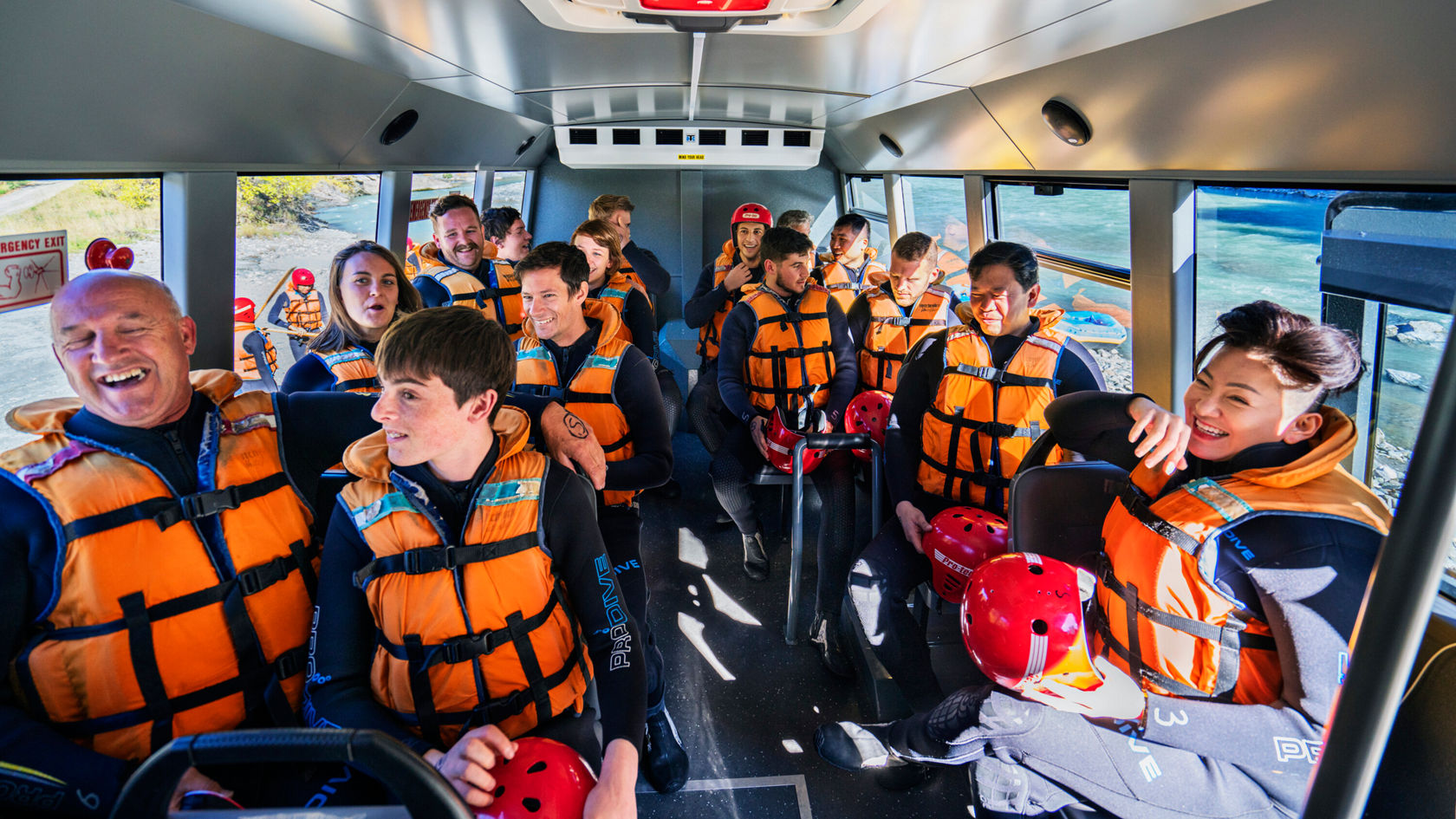 Eine Gruppe von Personen in orangefarbenen Schwimmwesten sitzt in einem Boot, lächelt und unterhält sich. Die Szene vermittelt eine Atmosphäre von Vorfreude auf ein Abenteuer im Freien.