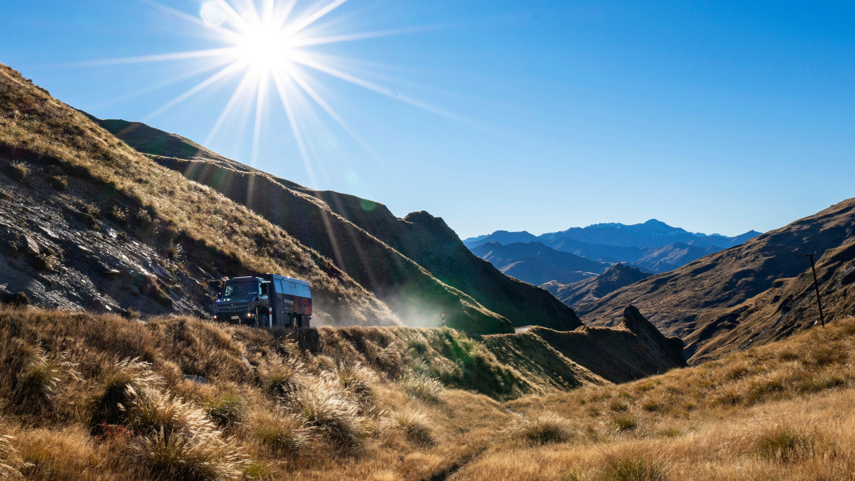 Ein Mercedes-Benz-Fahrzeug fährt durch eine bergige Landschaft mit sonnigem Himmel und beeindruckenden Bergen im Hintergrund, umgeben von grünem und trockenem Gras.