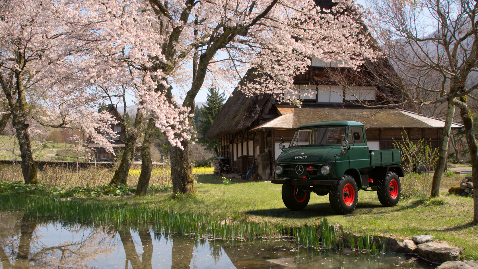 Een Mercedes-Benz Unimog staat op een weide naast een vijver, omgeven door bloeiende kersenbomen en met op de achtergrond een traditioneel vakwerkhuis.