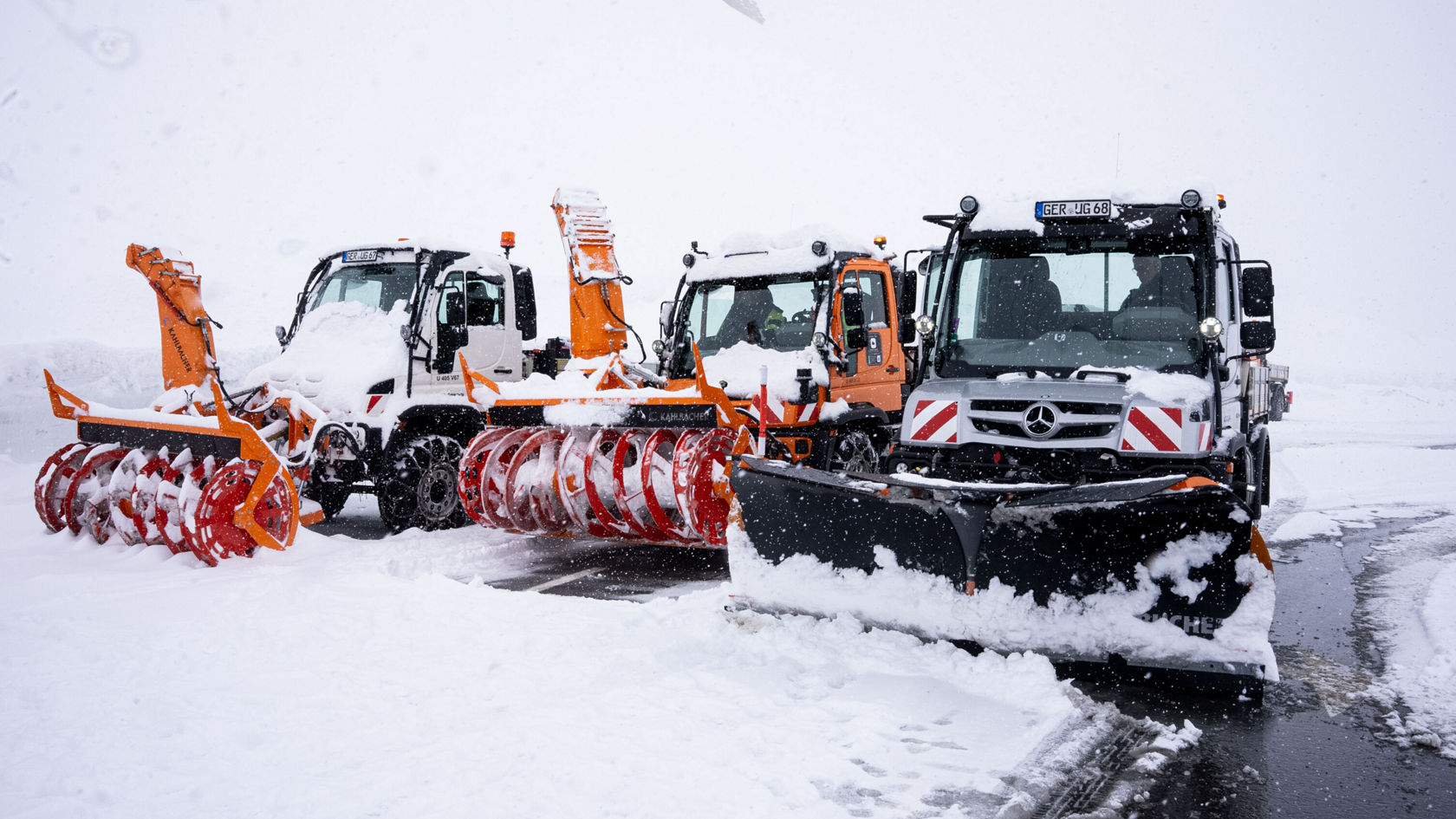Drei Schneeräumfahrzeuge, darunter ein Mercedes-Benz Unimog, stehen bei Schneefall im Schnee, ausgestattet mit Schneepflügen und -fräsen zur Winterdienstleistung.