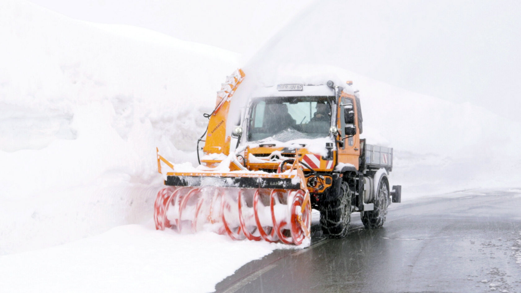 Ein Mercedes-Benz Unimog mit Schneepflug räumt eine schneebedeckte Straße frei. Starker Schneefall umgibt das Fahrzeug, das aktiv Schnee zur Seite schiebt.