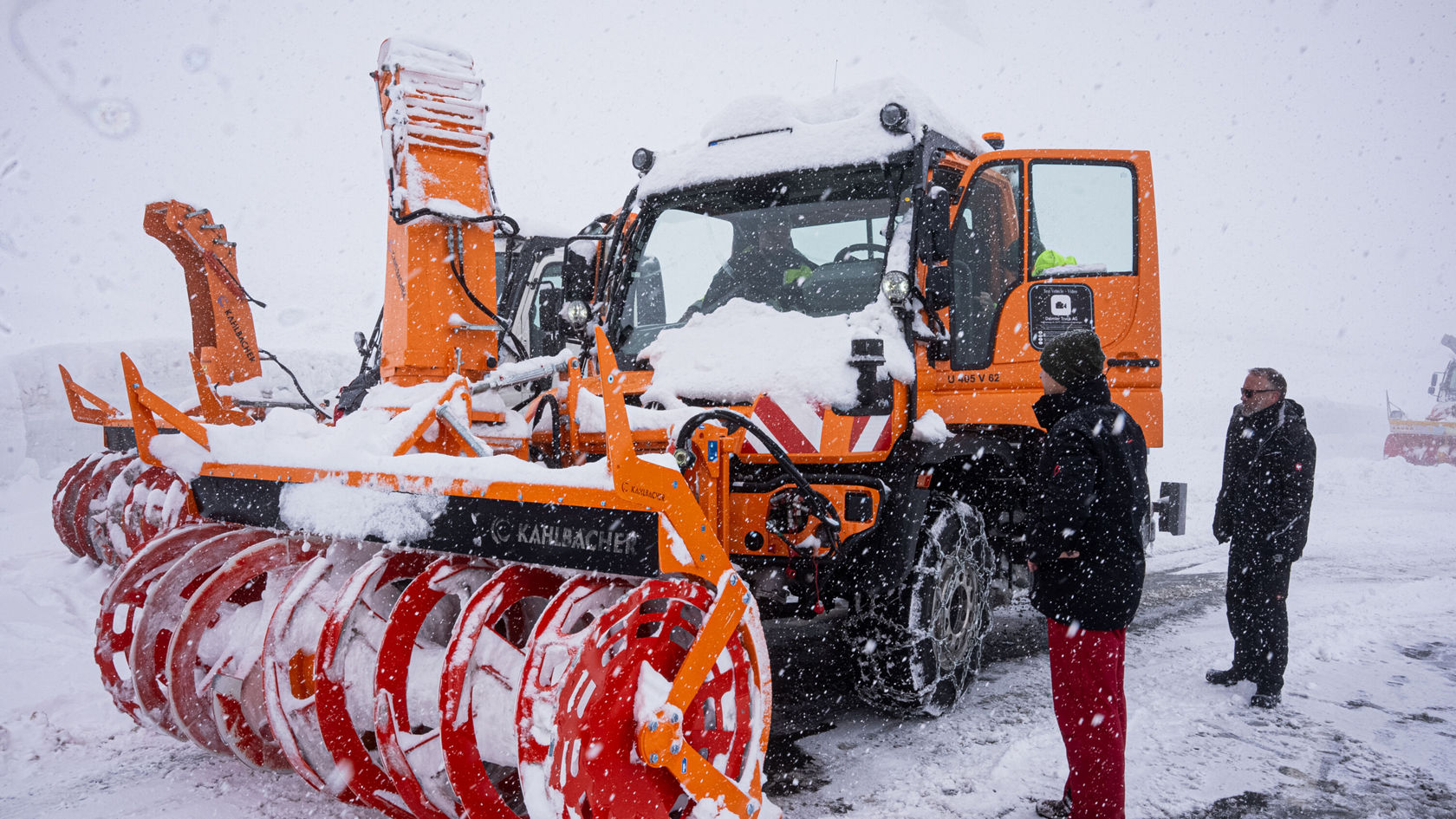 Ein Mercedes-Benz Unimog mit einem großen Schneepflug steht im Schnee, während zwei Personen daneben stehen und die Maschine beobachten.