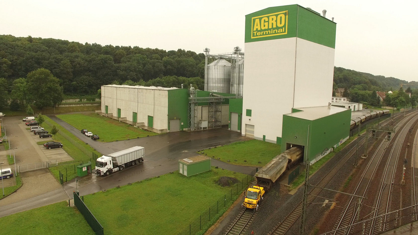Lagergebäude des Agro-Terminals mit hohen Silos und Lkw, die Waren liefern, neben einem Gleisbereich mit Zügen und Autobahn in einer grünen Umgebung.