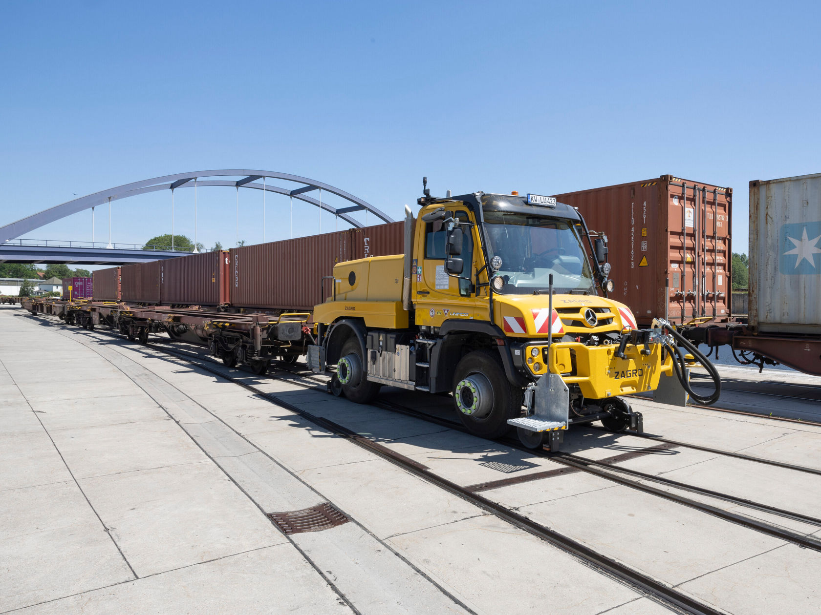 Ein gelber Unimog von Mercedes-Benz steht auf Schienen, neben mehreren Frachten in Containern. Der Unimog ist mit einem Kran ausgestattet, um Container zu verladen.