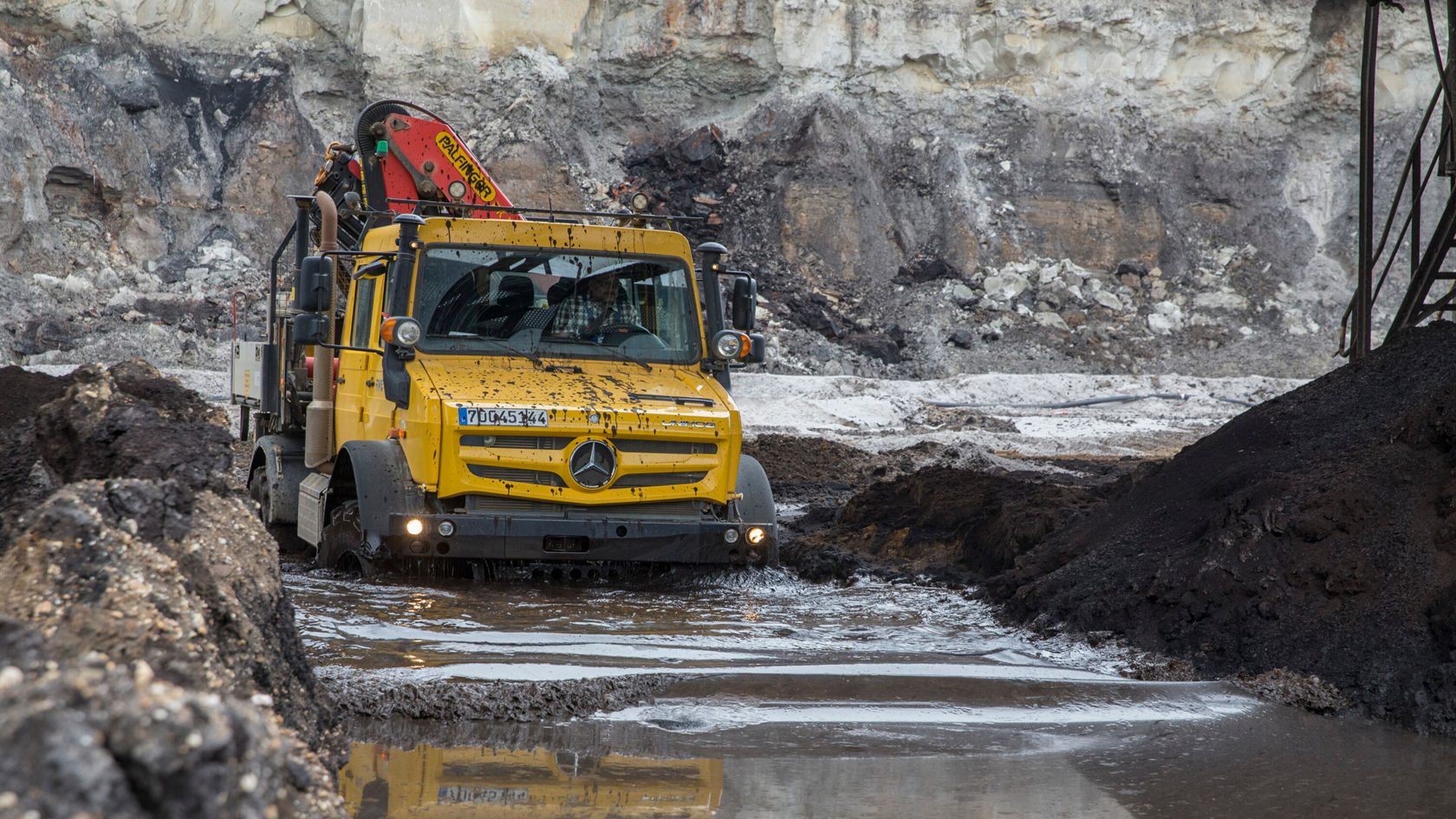 A yellow Mercedes‑Benz truck is driving through muddy terrain in a quarry, with a crane boom on the flatbed. The surroundings show geological strata and uneven ground.
