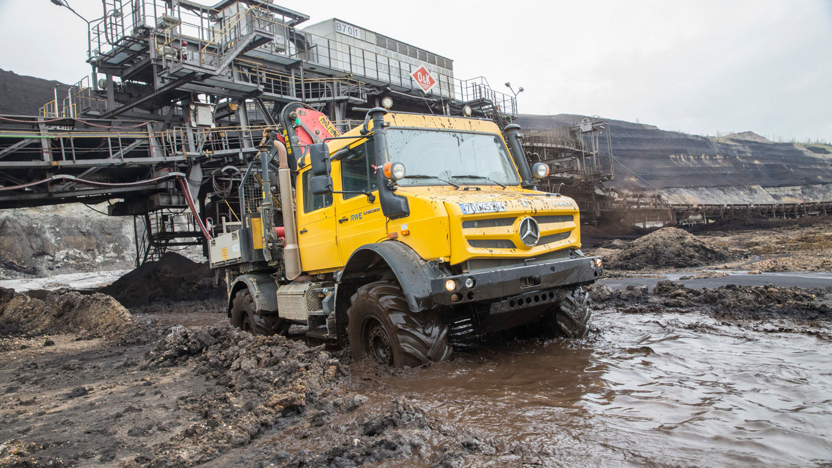 A yellow Mercedes-Benz Unimog drives through muddy terrain in an open-cast mine, with a conveyor system visible in the background.