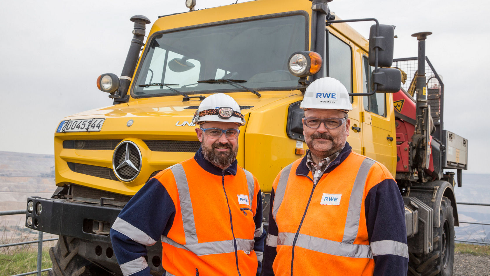 Two workers in safety clothing stand in front of a yellow Mercedes-Benz machine vehicle. They wear helmets and safety vests while they pose in an industrial environment.