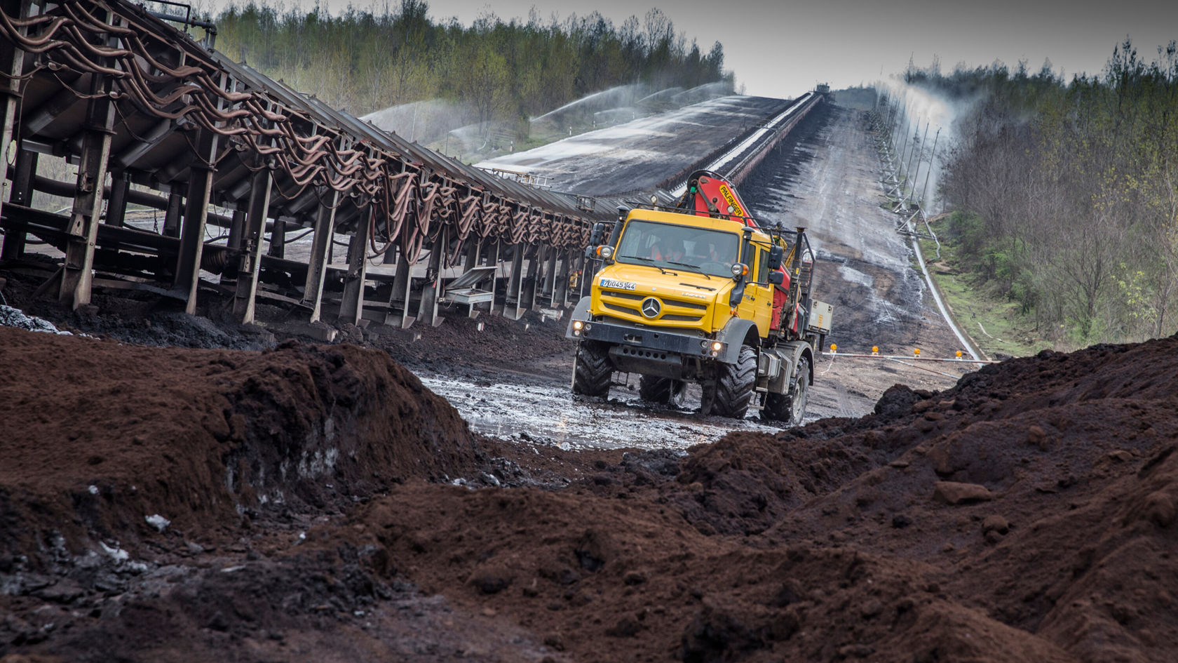 A yellow Mercedes-Benz Unimog is driving over uneven terrain in an industrial area, while a conveyor belt structure is visible in the background and steam is rising from the surroundings.