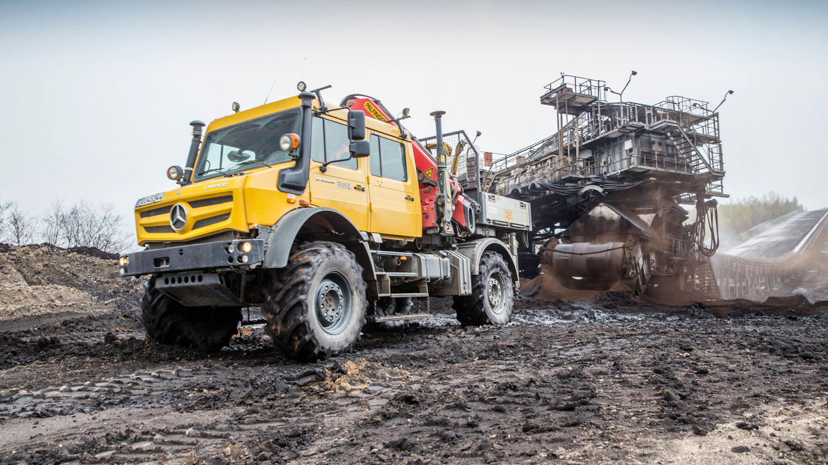 A yellow Mercedes-Benz Unimog is working on a construction site while standing near a large machine that is moving earth or material.
