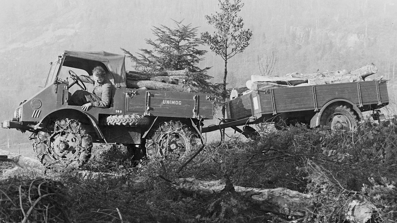 Ein Mercedes-Benz Unimog fährt über unwegsames Gelände, während ein Fahrer am Steuer sitzt und ein Anhänger mit Holzstämmen hinter sich zieht.