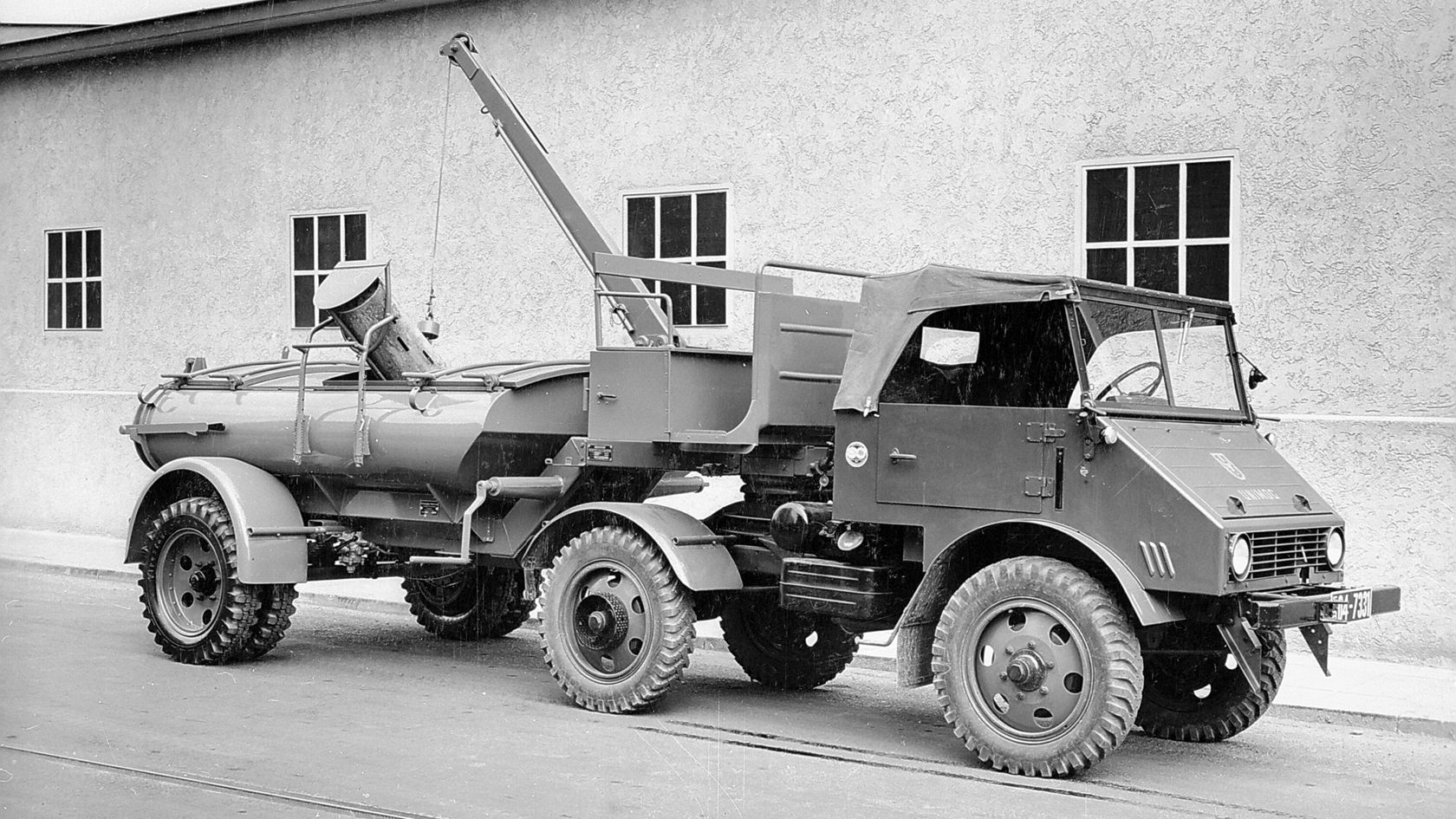 Ein Mercedes-Benz Unimog mit einem Kranaufbau steht auf einer Straße, daneben ein Mann in Uniform. Der Unimog wird in einem industriellen Umfeld gezeigt.