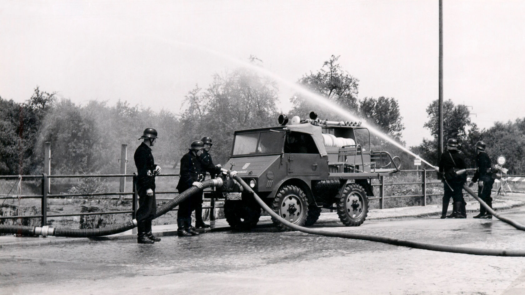 Ein Mercedes-Benz Unimog steht im Einsatz mit mehreren Feuerwehrleuten, die Schläuche bedienen und Wasser spritzen, um eine Löschübung durchzuführen.