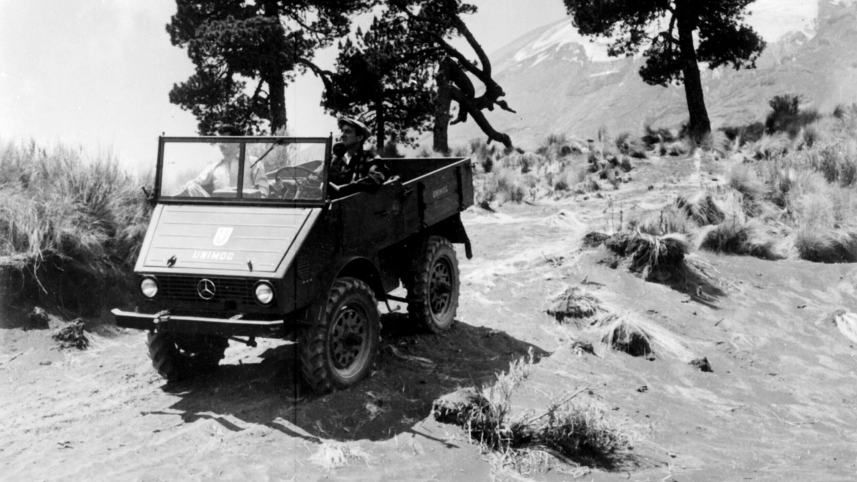 Ein Mercedes-Benz Unimog fährt durch unebenes Gelände, umgeben von Gras und Bäumen. Der Geländewagen zeigt eine robuste Bauweise und ist für Offroad-Abenteuer geeignet.