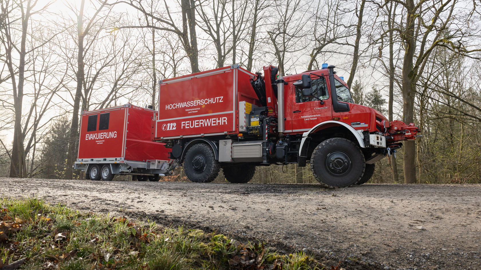 Un véhicule de pompiers Mercedes‑Benz rouge, avec une remorque portant l'inscription « protection contre les inondations » et « évacuation », est stationné sur un chemin entouré d'arbres.