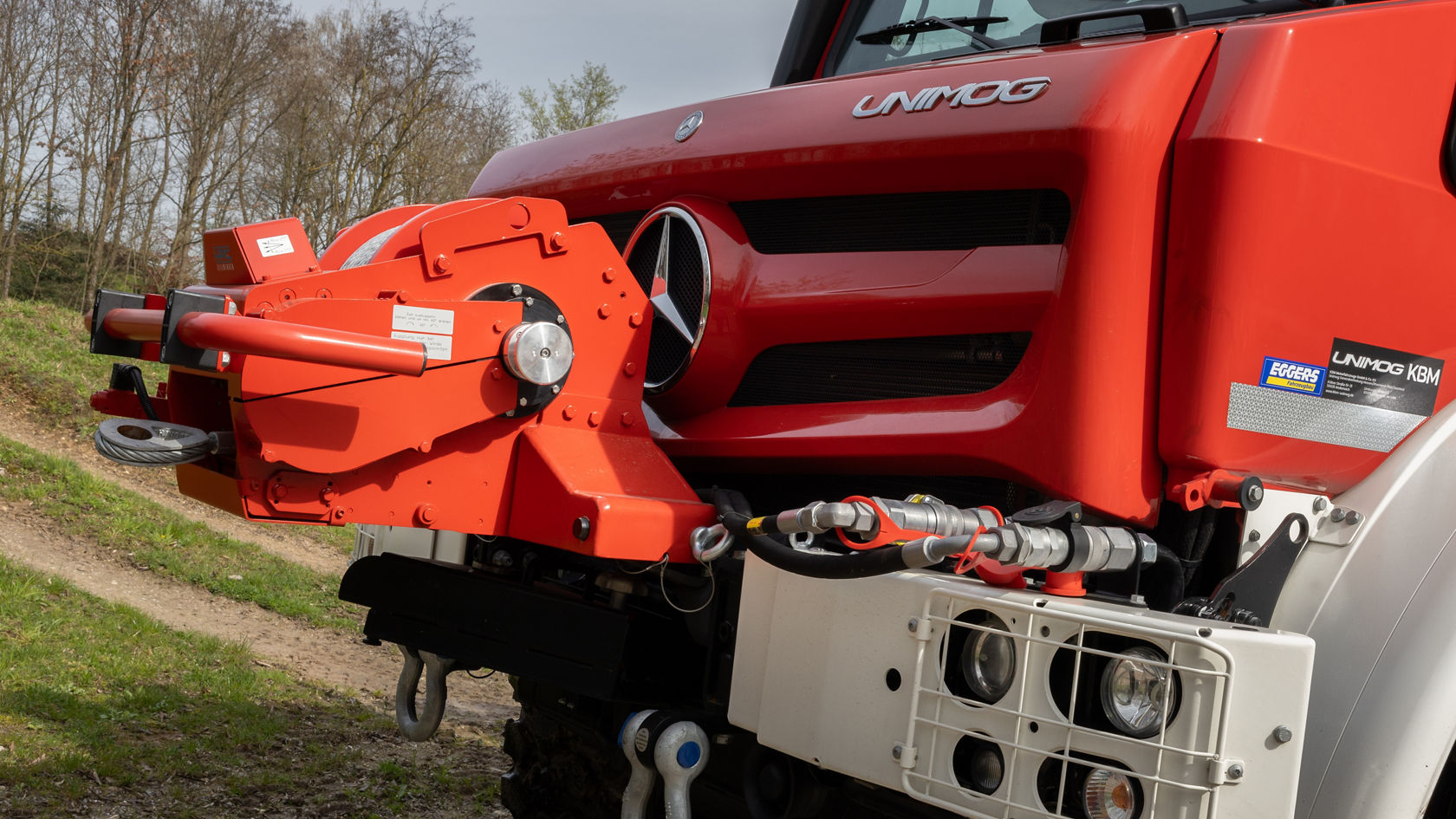 Un Unimog Mercedes‑Benz rouge équipé d'une plate-forme hydraulique à l'avant, préparé pour diverses applications tout-terrain, est stationné sur un chemin légèrement accidenté en milieu rural.