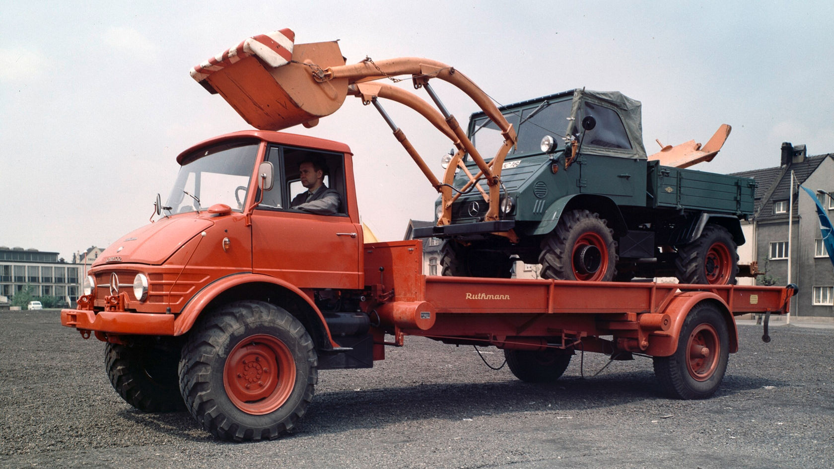 Rode Mercedes-Benz Unimog-transporter met een groene Mercedes-Benz Unimog op de laadbak, terwijl de graafarm van het rode voertuig omhoog wijst.