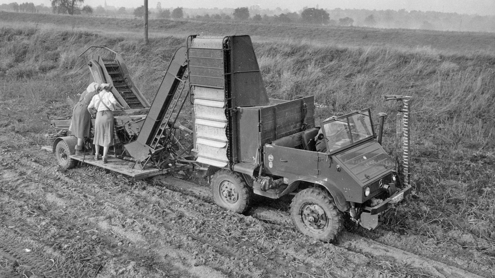 Unimog van Mercedes-Benz op een veld, gebruikt voor landbouwwerkzaamheden, met twee personen die aan een oogstmachine op de aanhanger werken.