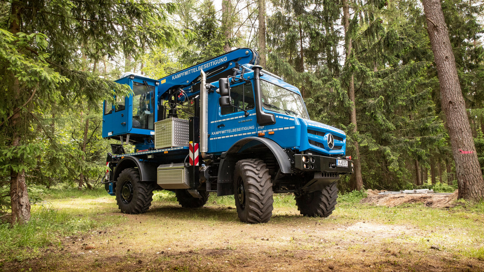 Un todoterreno Mercedes-Benz azul está sobre un suelo forestal, equipado con una grúa y neumáticos resistentes, rodeado de árboles altos y vegetación densa.