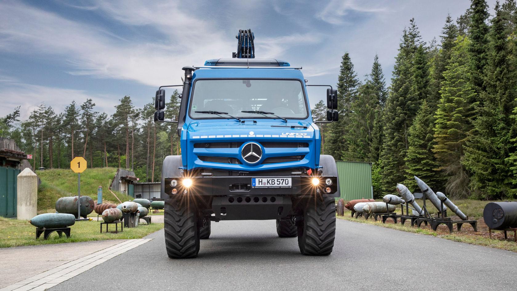 Un Mercedes-Benz Unimog azul está en una carretera, rodeado de árboles y de antiguos vehículos militares. Visionario y robusto, ideal para terrenos exigentes.