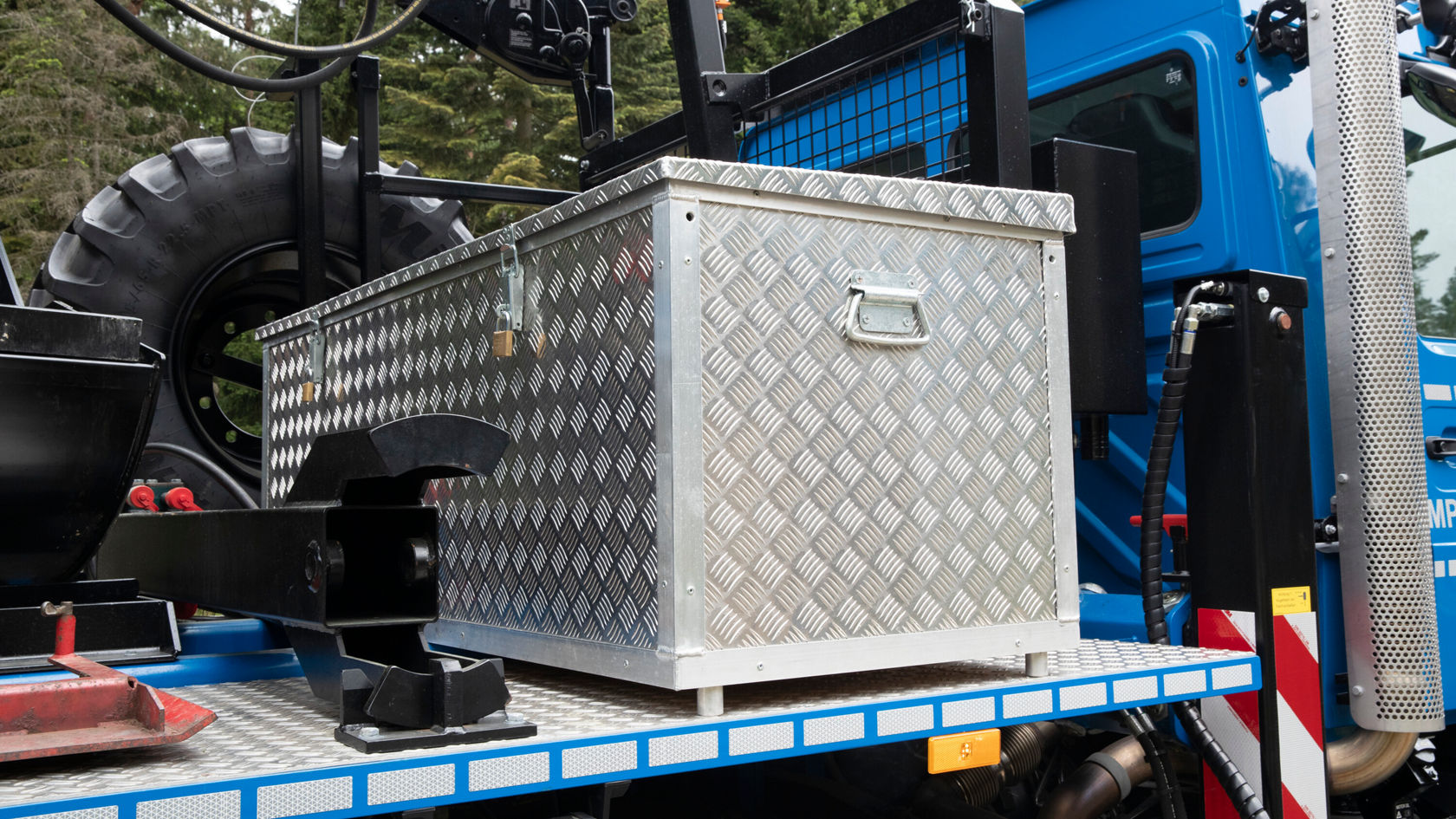 Una caja de transporte de aluminio plateada con patrón de rombos está sobre un vehículo. En el fondo se ven partes de un vehículo azul y una gran estructura de neumáticos.