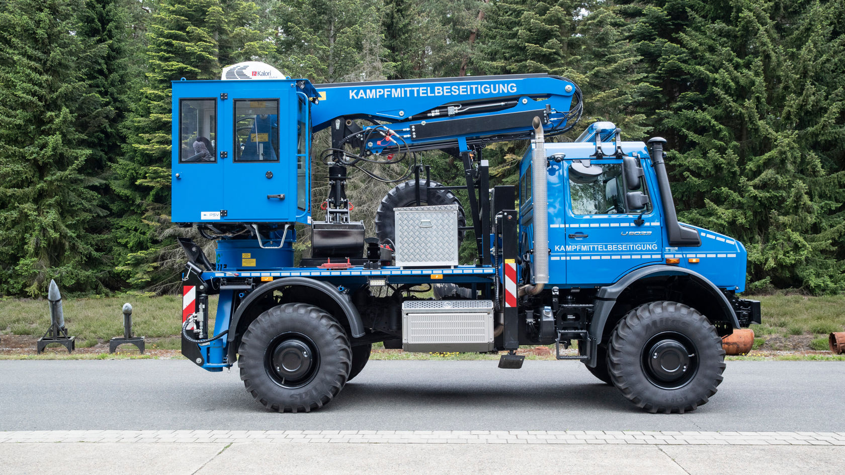 Un vehículo especializado azul para la desactivación de artefactos explosivos visto de perfil, equipado con una grúa y una cabina en la superestructura, frente a un paisaje boscoso.