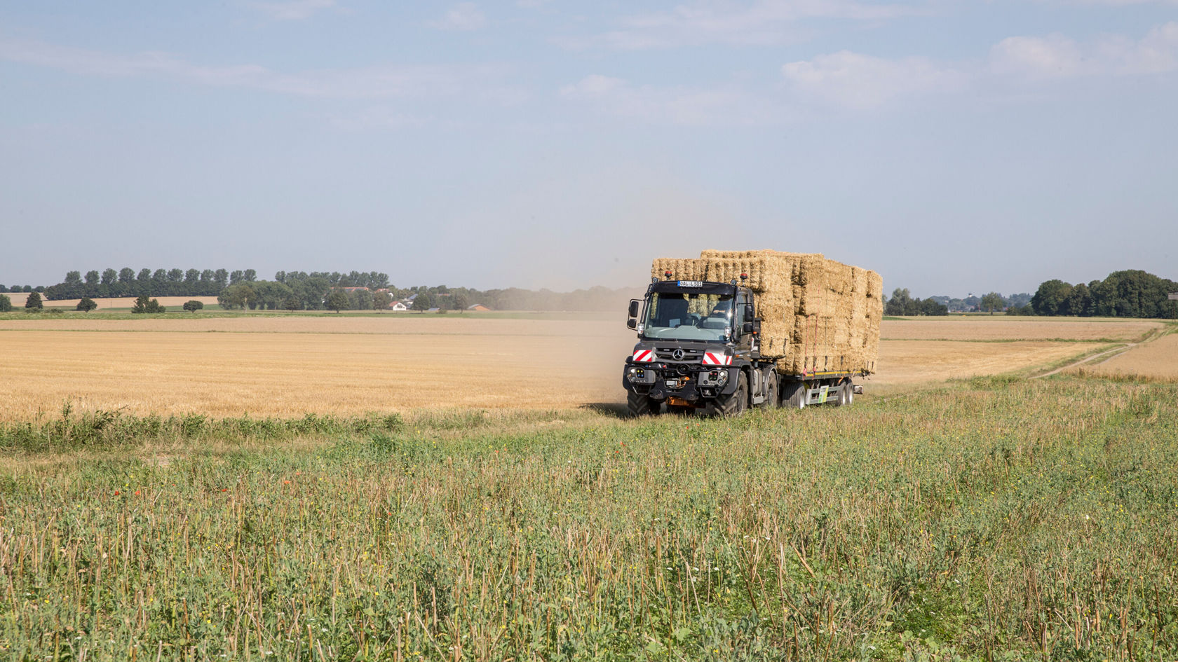 Un camion Mercedes‑Benz transporte des balles de foin à travers un champ, tandis que la poussière s'élève. À l'arrière-plan s'étendent des champs et des arbres sous un ciel bleu.