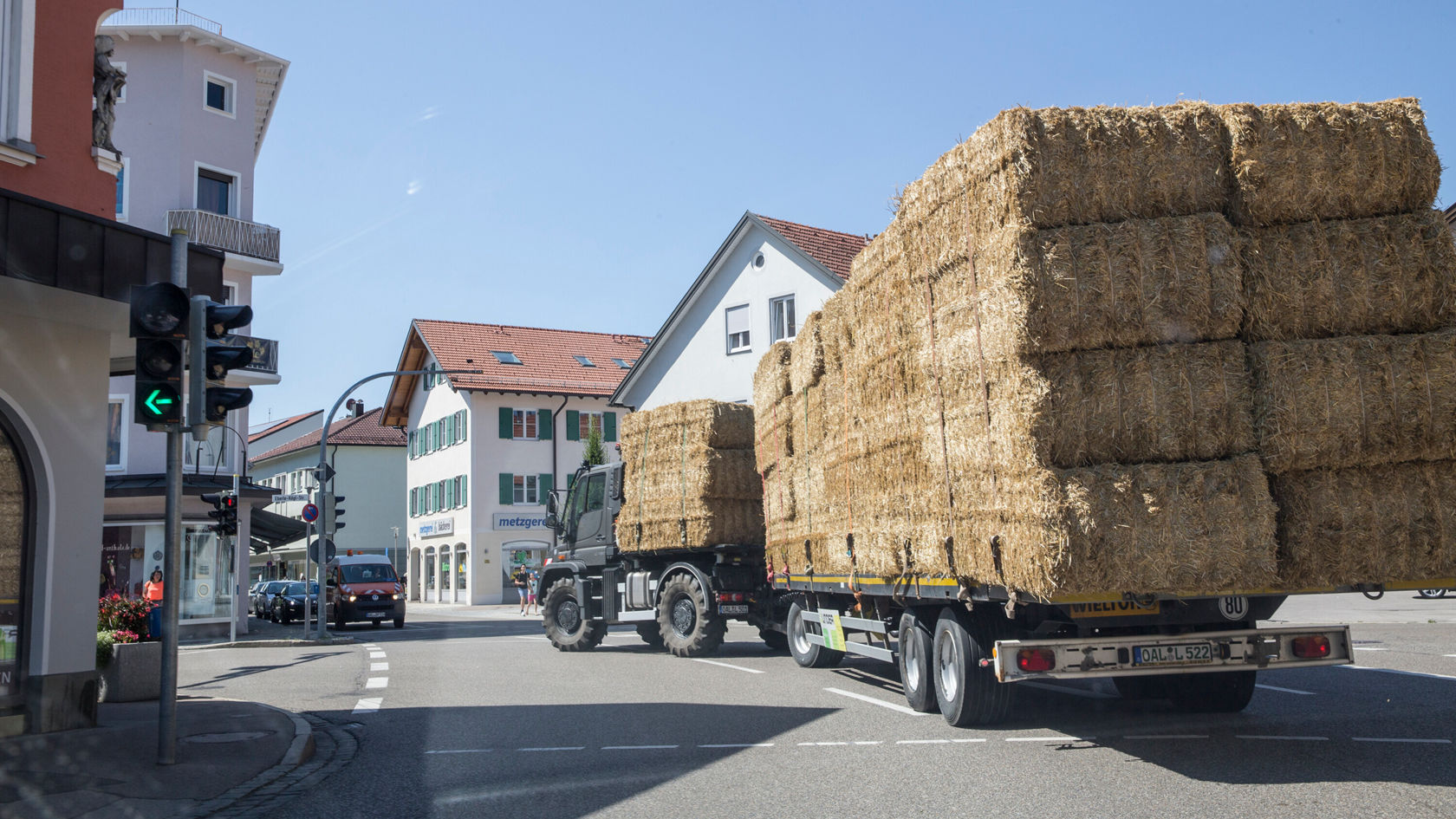 Un camion transporte des balles de paille en ville alors qu'il tourne à gauche à un feu. Des bâtiments sont visibles en arrière‑plan. Le temps est ensoleillé.