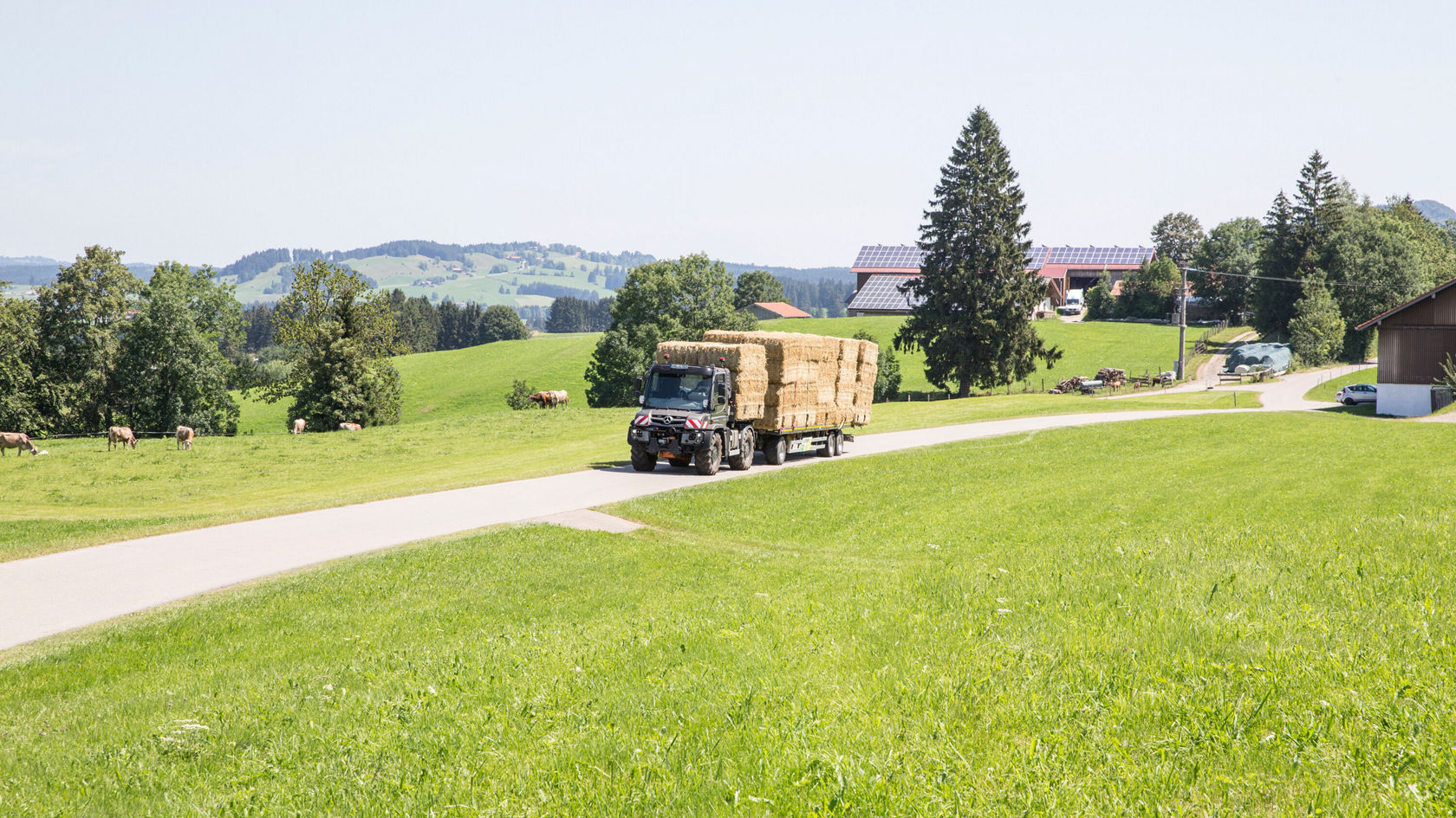 Un camion Mercedes-Benz transporte des balles de foin sur une route rurale. En arrière-plan, on aperçoit des prairies verdoyantes, des arbres et quelques bâtiments.