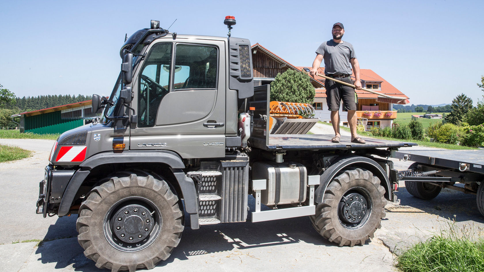 Un homme se tient sur la plate-forme de chargement d'un Unimog Mercedes‑Benz, alors qu'il tient un long bâton à la main. À l'arrière-plan, on aperçoit des bâtiments agricoles et des prairies verdoyantes.