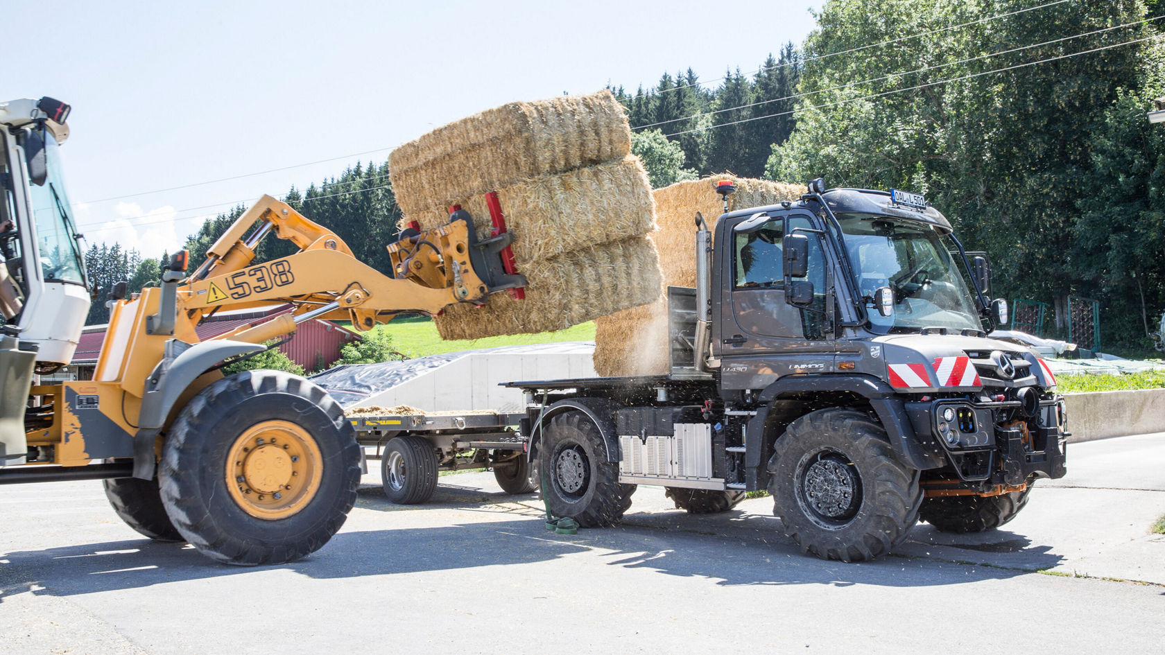 Un tracteur Mercedes‑Benz est en cours de chargement, tandis qu'une chargeuse sur pneus soulève des balles de foin et les charge sur une remorque. La scène se déroule dans un champ ou dans une cour.