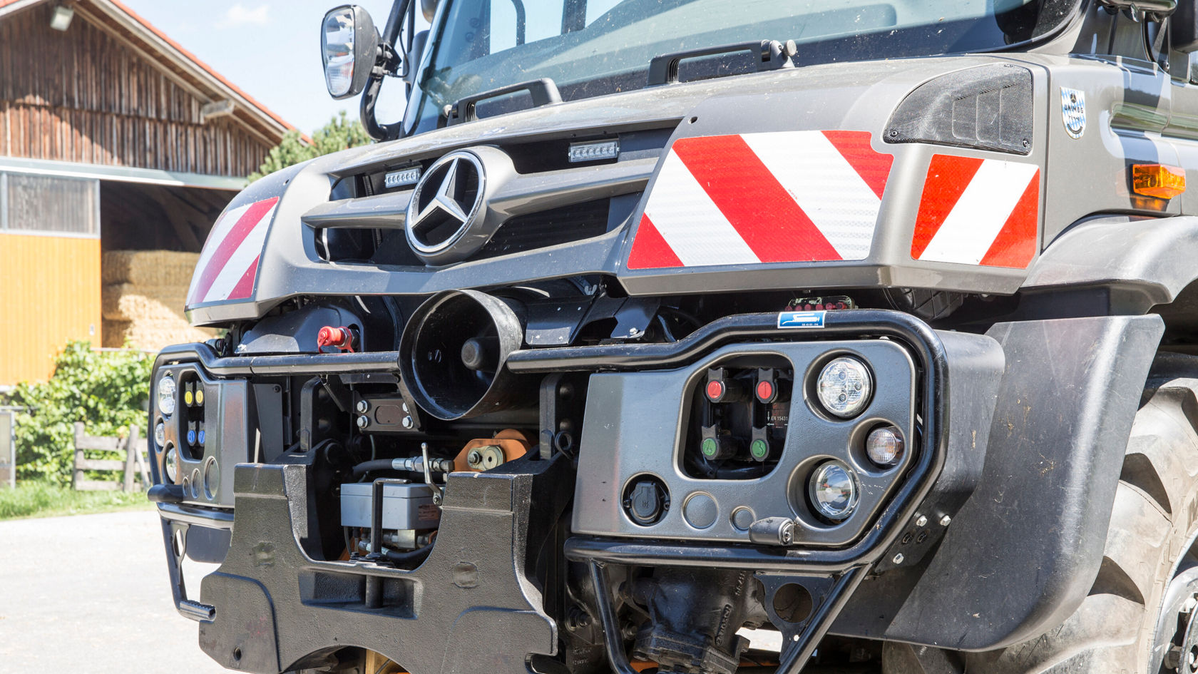 Vorderansicht des Stoßfängers eines Mercedes-Benz Unimog mit verschiedenen Kontrollleuchten, Schaltern und einem Anhängerkupplungssystem, geeignet für landwirtschaftliche Anwendungen.