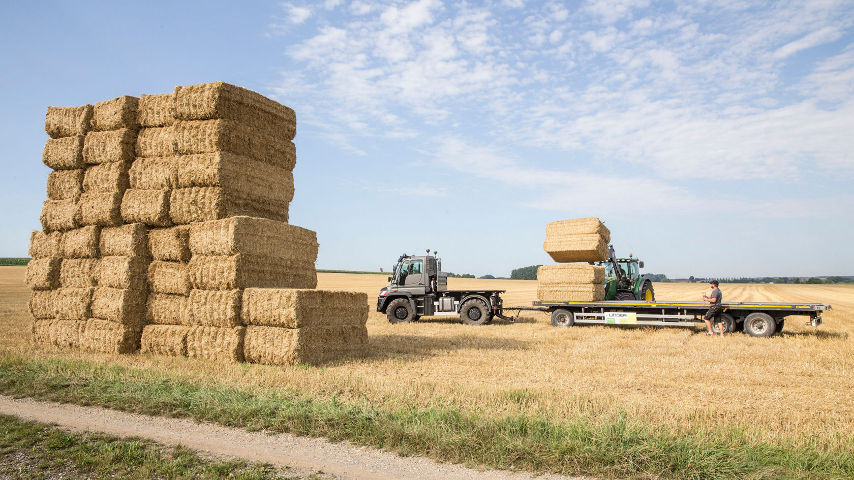 Des tracteurs transportent de grosses balles de foin sur un champ, tandis qu'une grande pile de balles de foin est visible en arrière‑plan. La scène montre des activités agricoles liées à la récolte.
