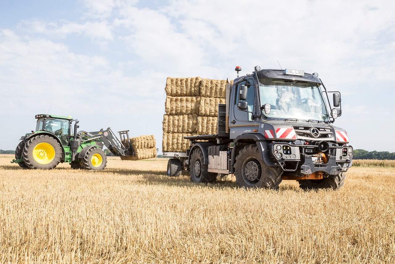 Un camion Mercedes‑Benz transporte des ballots de foin empilés dans un champ, tandis qu'un tracteur équipé d'un chargeur charge d'autres ballots de foin à bord du camion.