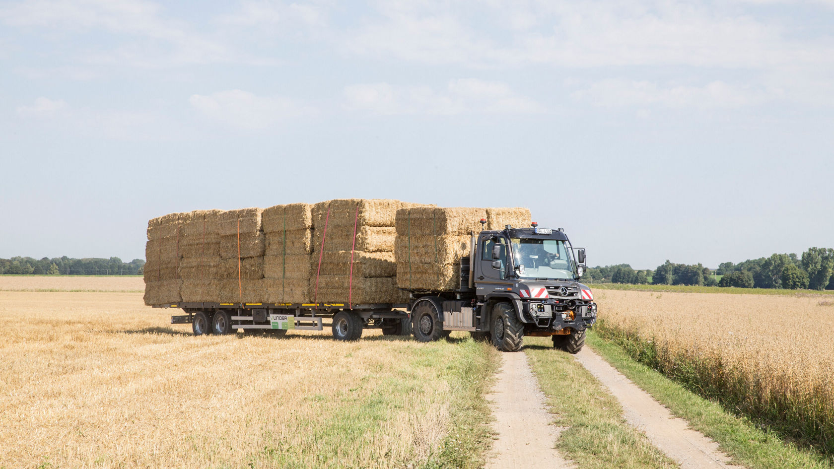 Un camion Mercedes‑Benz transporte de grandes balles de foin sur un chemin de terre, entouré d'un champ de céréales dorées, sous un ciel bleu.