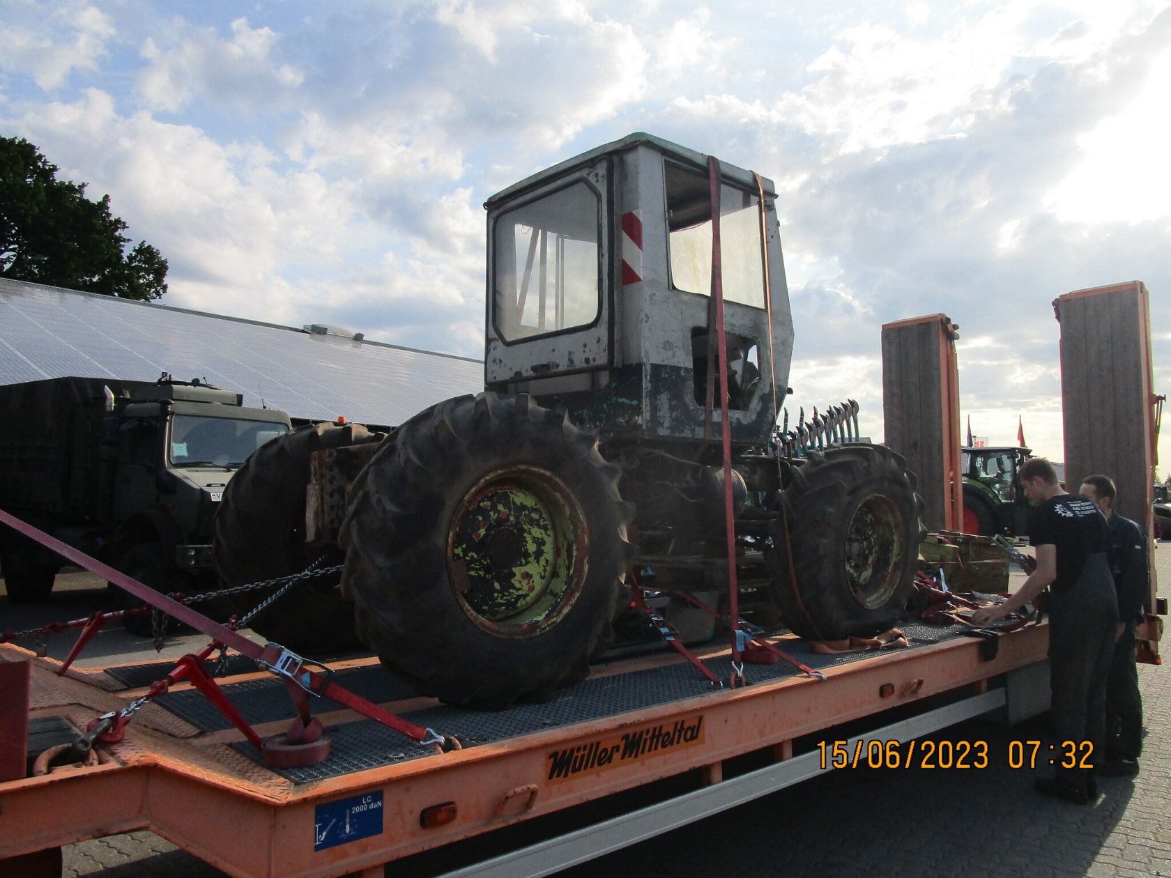 Ein schwerer Unimog von Mercedes-Benz steht auf einem Lkw-Anhänger, mit einem geschlossenen Fahrerschutzraum und großen, grobstolligen Reifen, während eine Person ihn sichert.