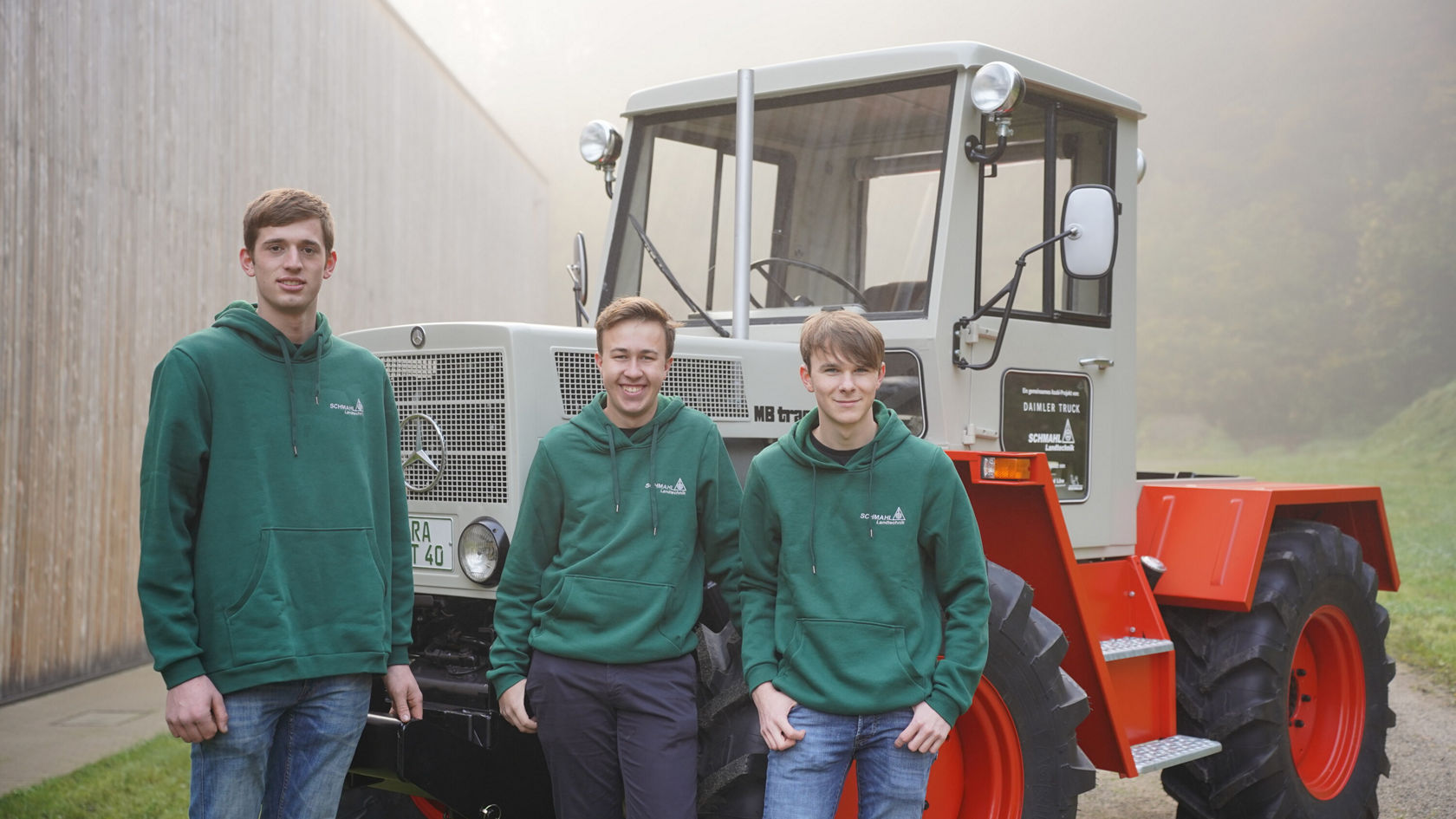 Drei junge Männer stehen vor einem Mercedes-Benz Unimog. Sie tragen grüne Kapuzenpullover und lächeln in die Kamera, während der Unimog teilweise verdeckt von Nebel ist.