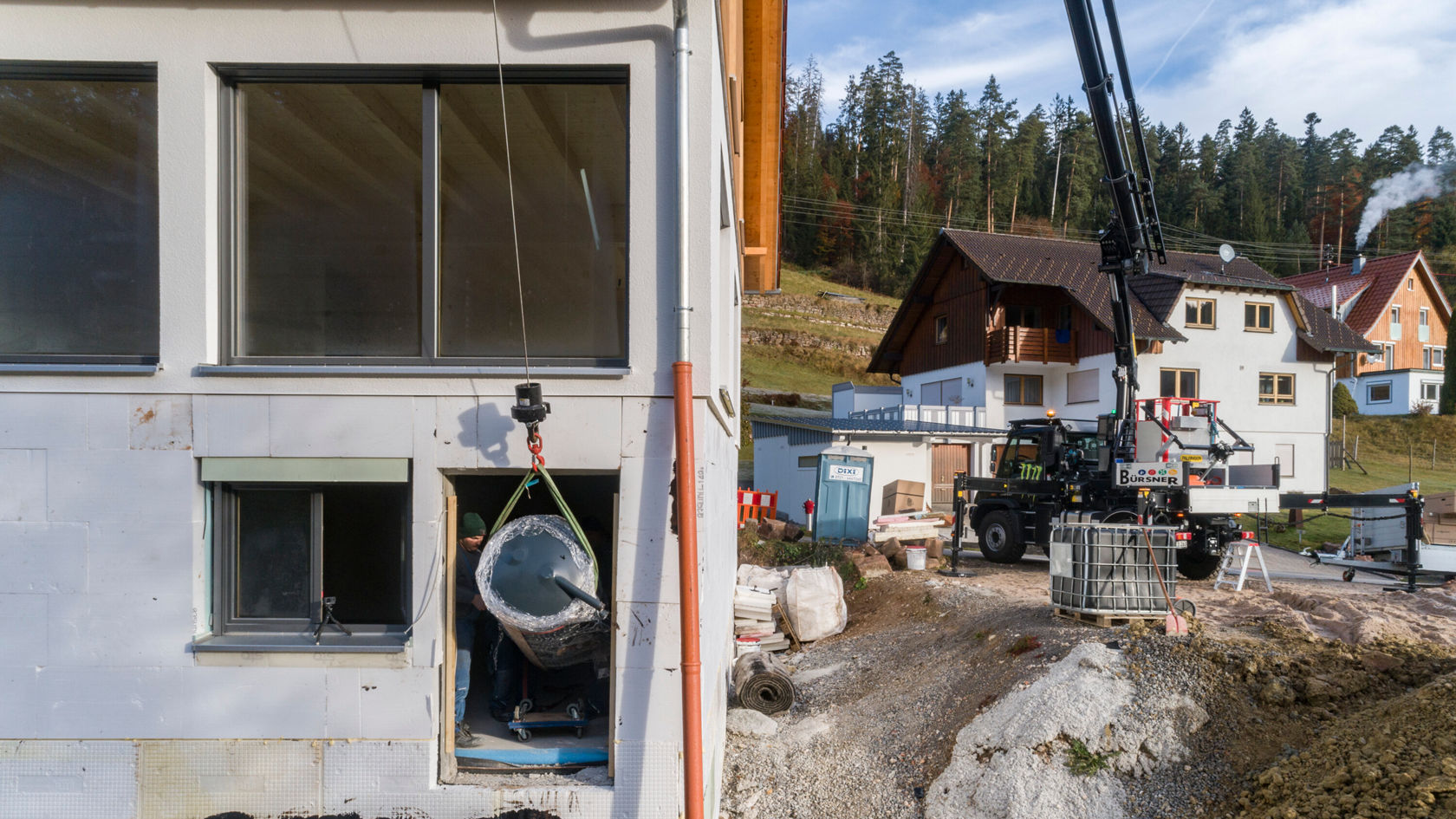 Ein Kran hebt einen großen Gegenstand in der Nähe eines Wohnhauses, umgeben von Baustellenmaterialien und weiteren Gebäuden im Hintergrund, in einer bewaldeten Landschaft.