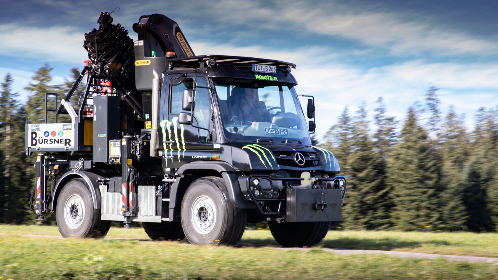 Robuster Mercedes-Benz Unimog in Schwarz, ausgestattet mit einem kranähnlichen Aufbau, fährt durch einen Wald. Auf der Seite ist ein Logo des Sponsors sichtbar.
