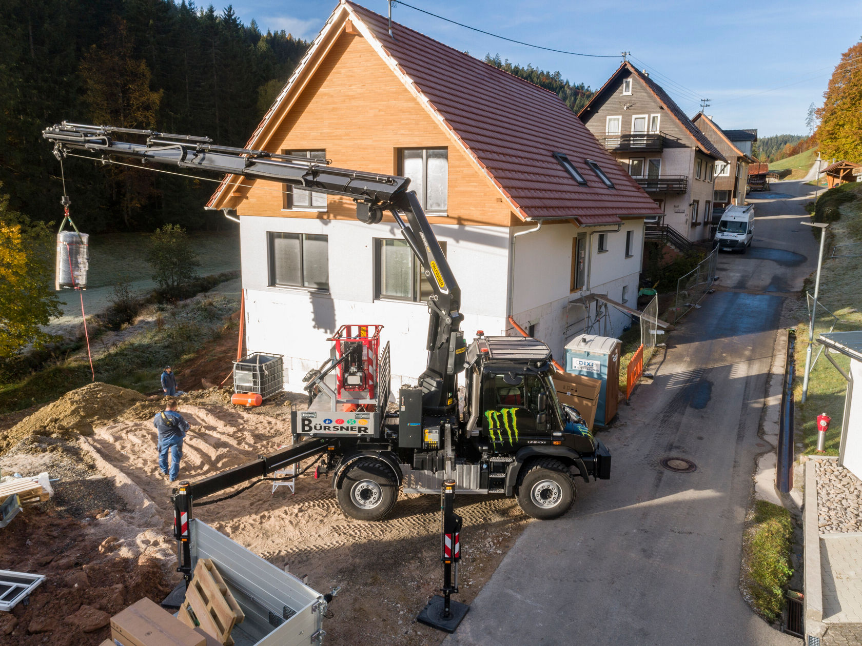 Un camion Mercedes‑Benz équipé d'une grue soulève une charge de chantier dans un quartier résidentiel. Un ouvrier se tient à côté du véhicule, tandis qu'une maison est visible en arrière-plan.