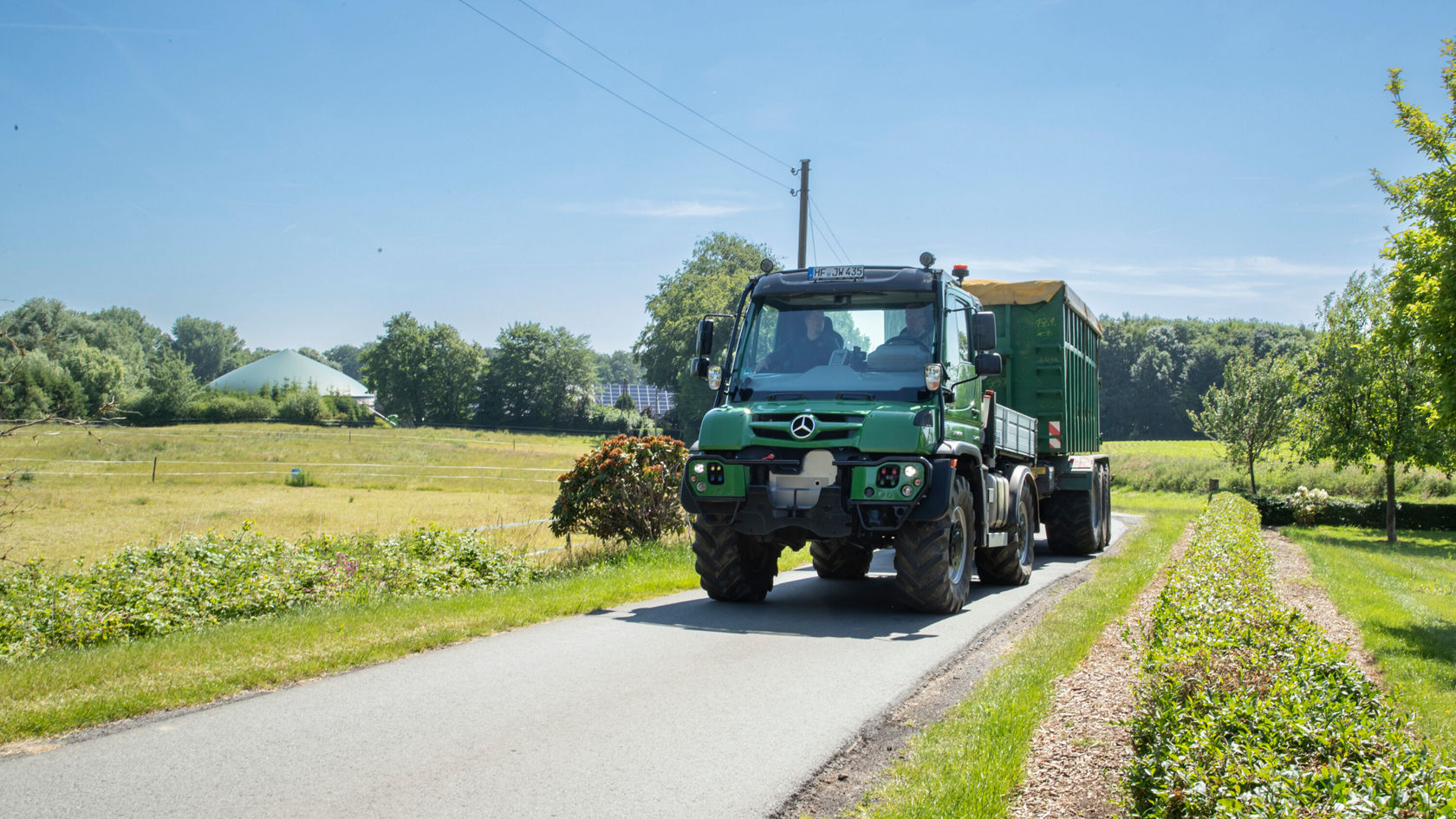 Ein grüner Mercedes-Benz Lkw fährt auf einer Landstraße vorbei an Feldern und Bäumen, mit einem Auflieger, der mit einer Plane bedeckt ist.