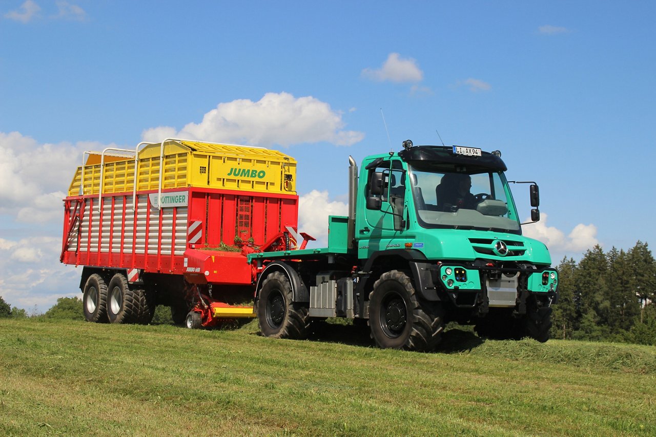 Ein grüner Mercedes-Benz Unimog zieht einen roten Anhänger mit gelbem Aufsatz, der mit landwirtschaftlichem Material beladen ist, auf einer Wiese unter blauen Himmel.