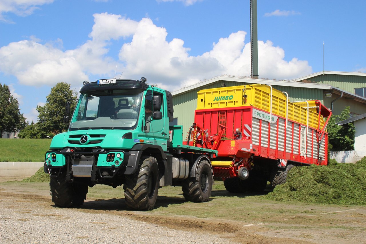 Ein grüner Mercedes-Benz Unimog steht auf einem Feld neben einem roten, mit gelbem Aufsatz versehenen Anhänger zur Ernte.