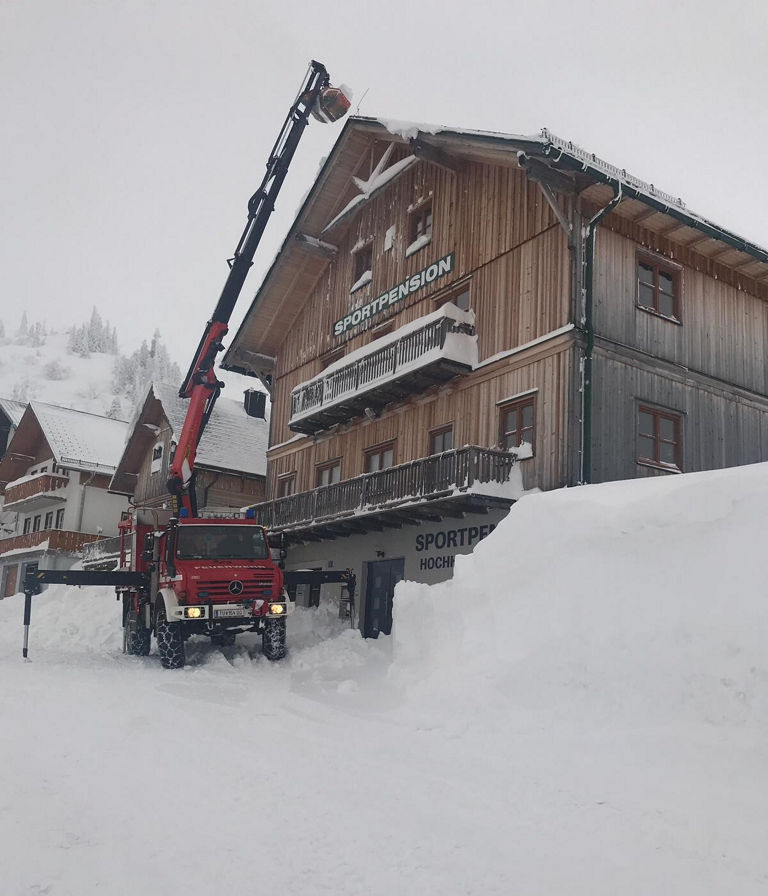 Ein Mercedes-Benz Lkw mit einem Kran hebt Materialien zur Bergung von Schnee an einem verschneiten Gebäude in den Bergen. Der Eingang zum Hotel "Sportpen" ist stark eingeschneit.