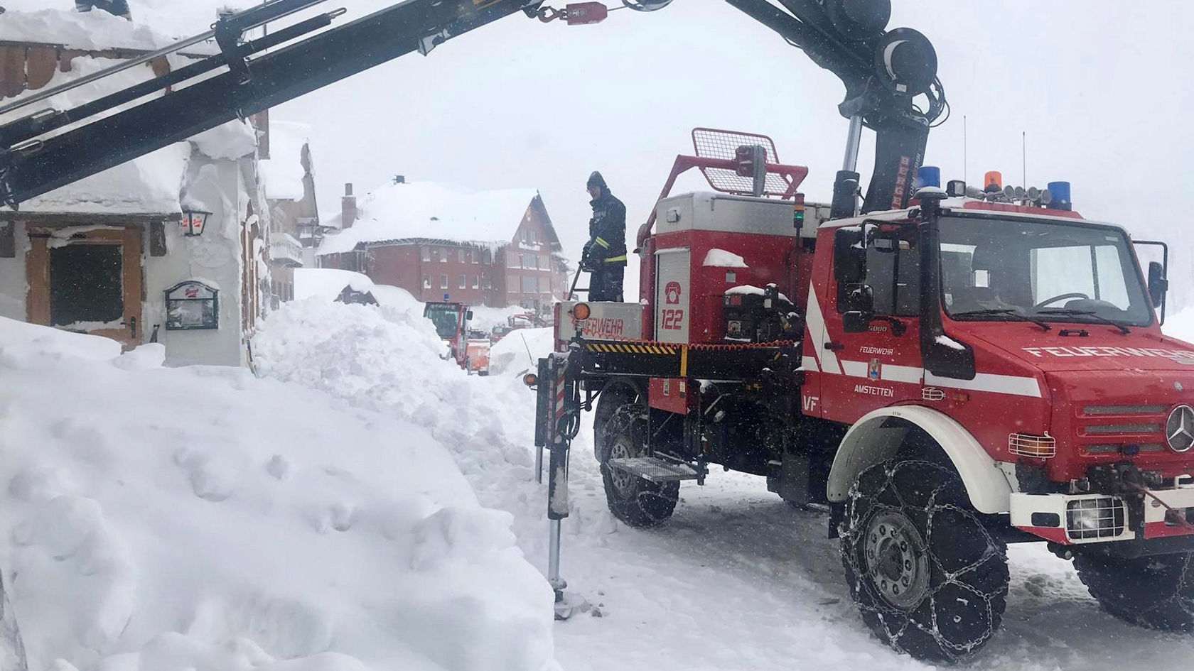 Ein Mercedes-Benz Einsatzfahrzeug steht neben einem schneebedeckten Gebäude. Ein Arbeiter nutzt ein Gerät zur Schneebeseitigung in einer winterlichen Umgebung.