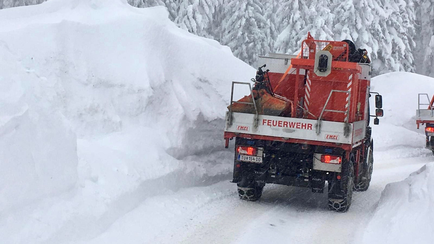Ein Feuerwehrfahrzeug mit Schneepflug fährt durch eine verschneite Landschaft, um Straßen von hohen Schneeansammlungen zu räumen. Die Umgebungsbedingungen sind winterlich und schneebedeckt.