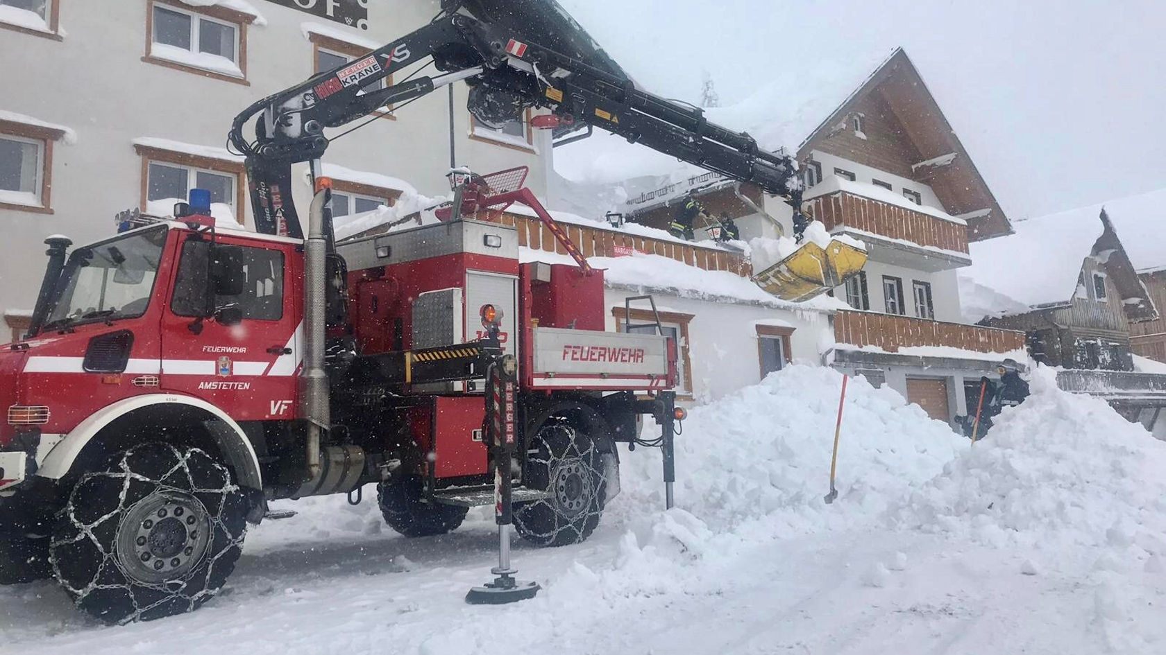 Ein Mercedes-Benz Feuerwehrfahrzeug mit einem Kran entfernt Schnee von einem Balkon während eines Schneesturms. Hohe Schneehaufen sind im Vordergrund zu sehen.