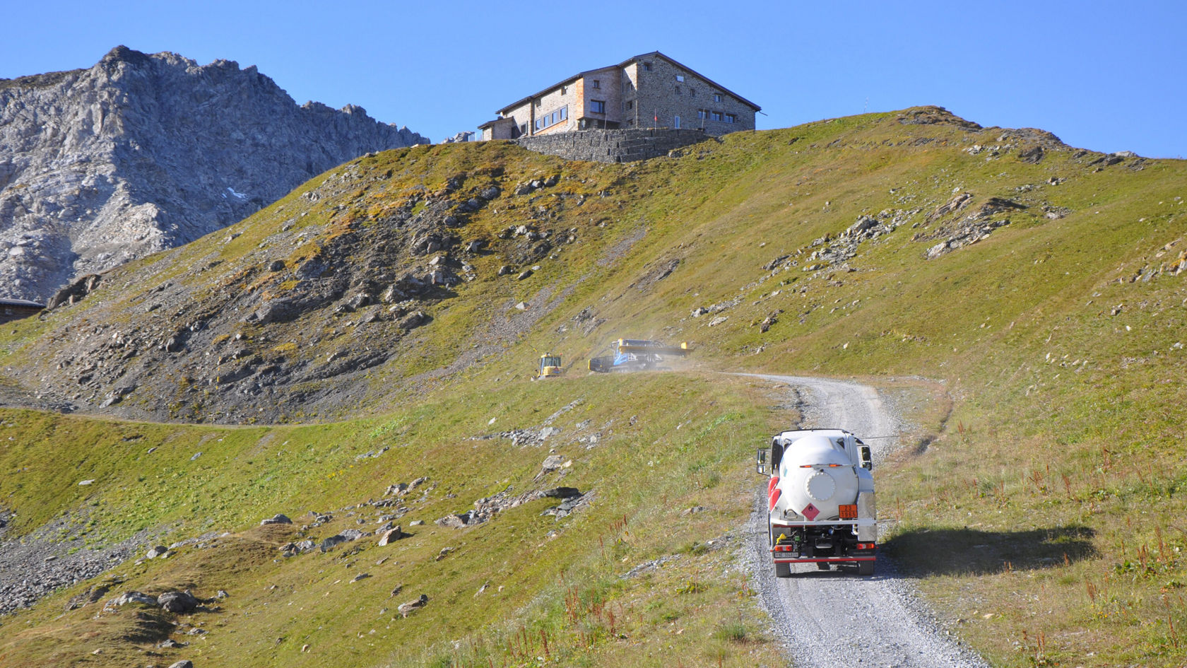 Ein Mercedes-Benz Fahrzeug fährt eine kurvenreiche Schotterstraße hinauf zu einem Berghaus, umgeben von grünen Wiesen und Bergen im Hintergrund.