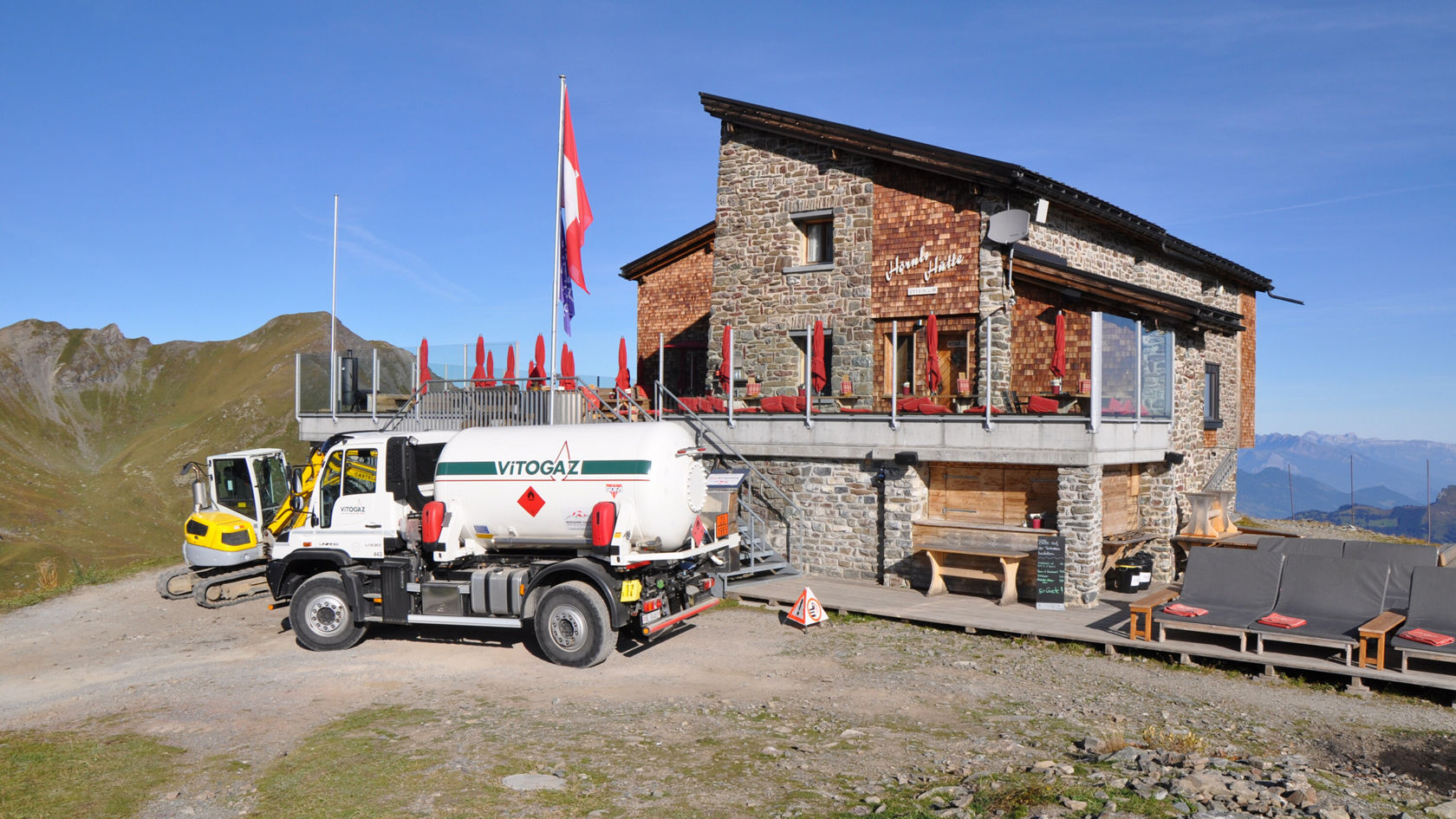 Ein Berggasthaus aus Stein mit einer großen Fensterfront und rot dekorierten Tischen im Freien. Ein Lastwagen mit Gaszellen steht davor.