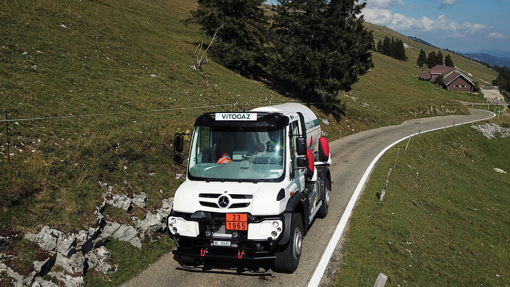Mercedes-Benz-LKW auf einer kurvenreichen Straße in einer ländlichen Umgebung, mit einer Tankstelle im Hintergrund, transportiert möglicherweise Flüssiggas.